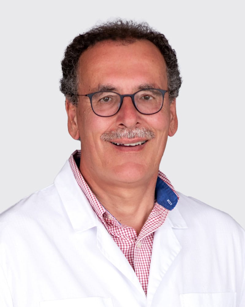 Arzt in weißem Kittel mit Brille, lächelnd, vor hellem Hintergrund, professionell und vertrauenswürdig, repräsentiert medizinische Kompetenz und Patientenversorgung.